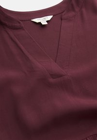 Burgundy v-kauluspaita sileässä kankaassa, jossa on pleatattu yksityiskohta, hienovaraiset tikkaukset kauluksessa ja lyhyet hihat.