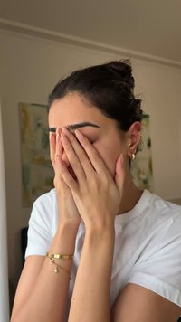 Donna con camicia bianca che si copre il viso con entrambe le mani, indossando braccialetti d'oro e diversi orecchini, all'interno con sfondo sfocato.