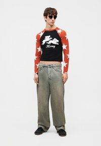 Mladý muž nosiaci čierny a červený dlhý crop top s hviezdičkovým vzorom, bielymi zajacmi a nápisom "Horny", voľné sivé džínsy, čierne topánky a slnečné okuliare.