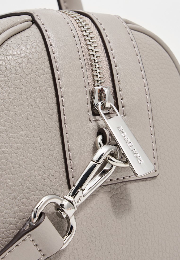 Sac en cuir texturé gris avec une fermeture éclair en métal et des accessoires en acier, accompagné d'une étiquette suspendue affichant le nom de la marque.