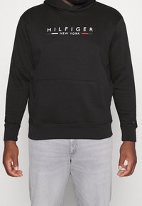 Felpa nera con cappuccio e tasca frontale, con il logo "HILFIGER NEW YORK" in bianco, rosso e blu. Tessuto morbido e vestibilità standard.