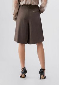 Shorts marron sur mesure avec une finition texturée et une unique poche arrière. Associés à des talons noirs à lanières. Petit pli subtil à l'avant.
