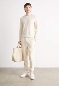 Beige longsleeve shirt en bijpassende joggers van zachte stof. Draagt een lichtbeige schoudertas met twee handvatten. Witte sneakers.