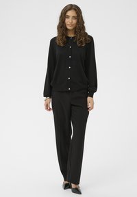 Fekete, gombos blúz texturált felülettel, elülső gombokkal és hosszú ujjakkal, fekete szabott nadrággal és hegyes orrú fekete cipővel párosítva.