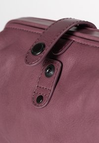Dettaglio di una borsa in pelle viola con una patta e chiusura a pressione nera, e cuciture a contrasto lungo i bordi. Texture liscia visibile.