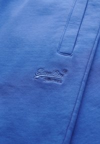 Tela azul con un logo SuperDry bordado y un bolsillo con abertura vertical visible cerca de la esquina superior derecha.