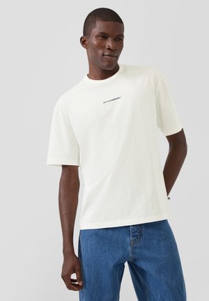 Młody mężczyzna ubrany w białą koszulkę z krótkim rękawem z napisem "LET’S SYNERGIZE!" oraz niebieskie dżinsy, stoi na tle jednolitego jasnego tła.