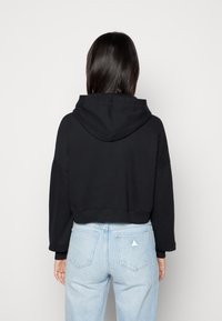 Felpa cropped nera con cappuccio, maniche lunghe e orlo a coste, abbinata a jeans azzurri chiari con una toppa triangolare sulla tasca posteriore.