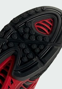 Nahaufnahme einer schwarz-roten Schuhsohle mit runden Grip-Noppen und strukturierten Profilmustern für besseren Halt.