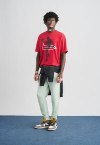 Κόκκινο υπερμεγέθες μπλουζάκι με γραφικό μπάσκετ, σε συνδυασμό με τζόγκερ σε μέντα πράσινο και ψηλά αθλητικά παπούτσια σε γκρι και μαύρο. Μαύρο πουλόβερ δεμένο στη μέση.