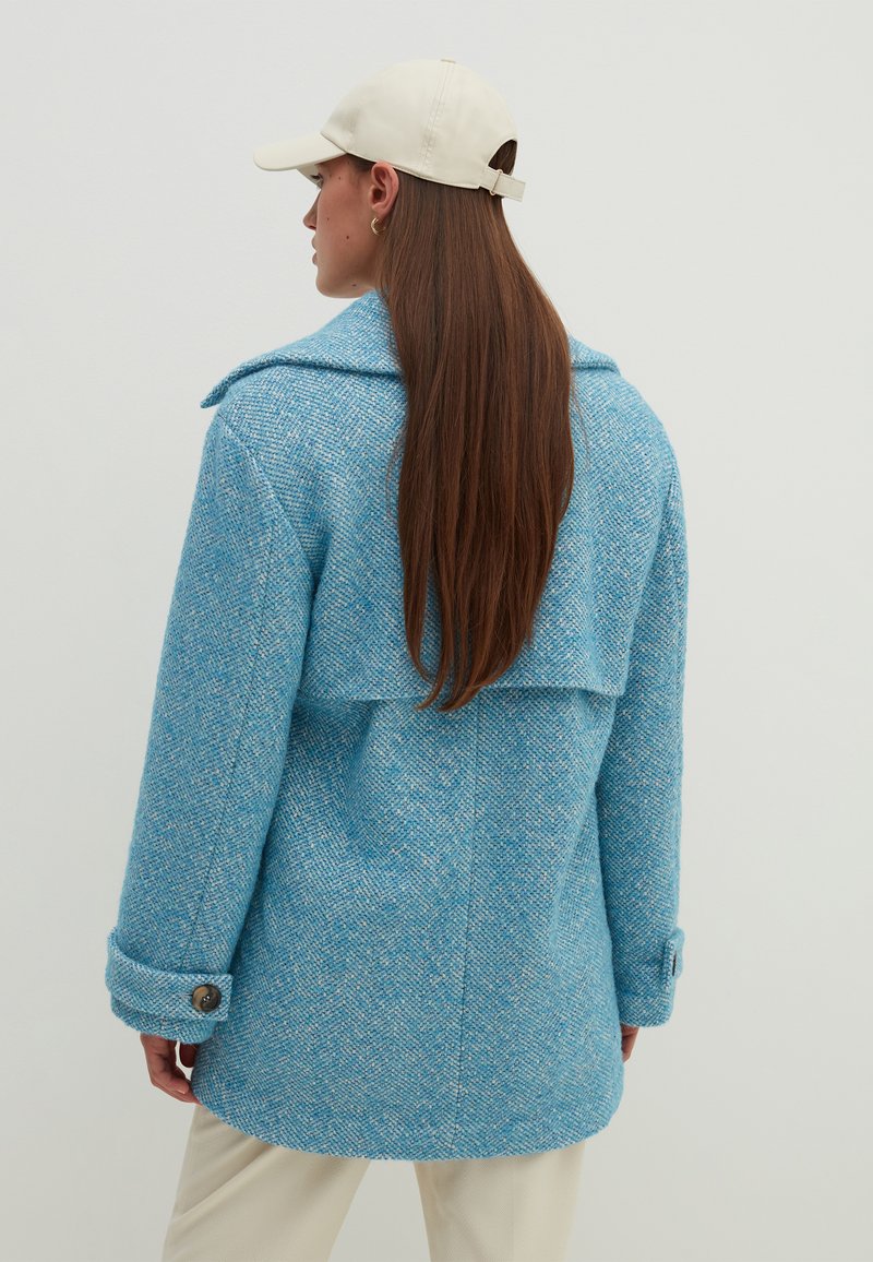 Blauer Tweedmantel mit breitem Kragen, geknöpften Ärmelbündchen und geschichteter Rückengestaltung. Getragen mit einer hellen Mütze. Glatte Textur.