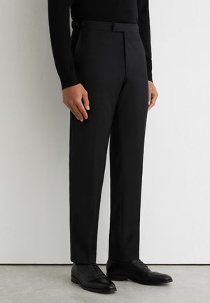 Man draagt op maat gemaakte zwarte nette broek, zwarte top met lange mouwen en zwarte leren nette schoenen, staand binnenshuis.