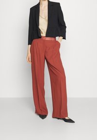 Un mannequin se tient en portant un blazer noir court, un haut beige et un pantalon large couleur rouille, adoptant une pose décontractée avec les mains dans les poches.