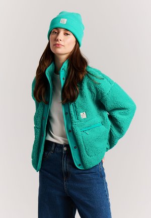 Woman wearing teal fleece jacket and matching beanie with white shirt and blue jeans against plain background.