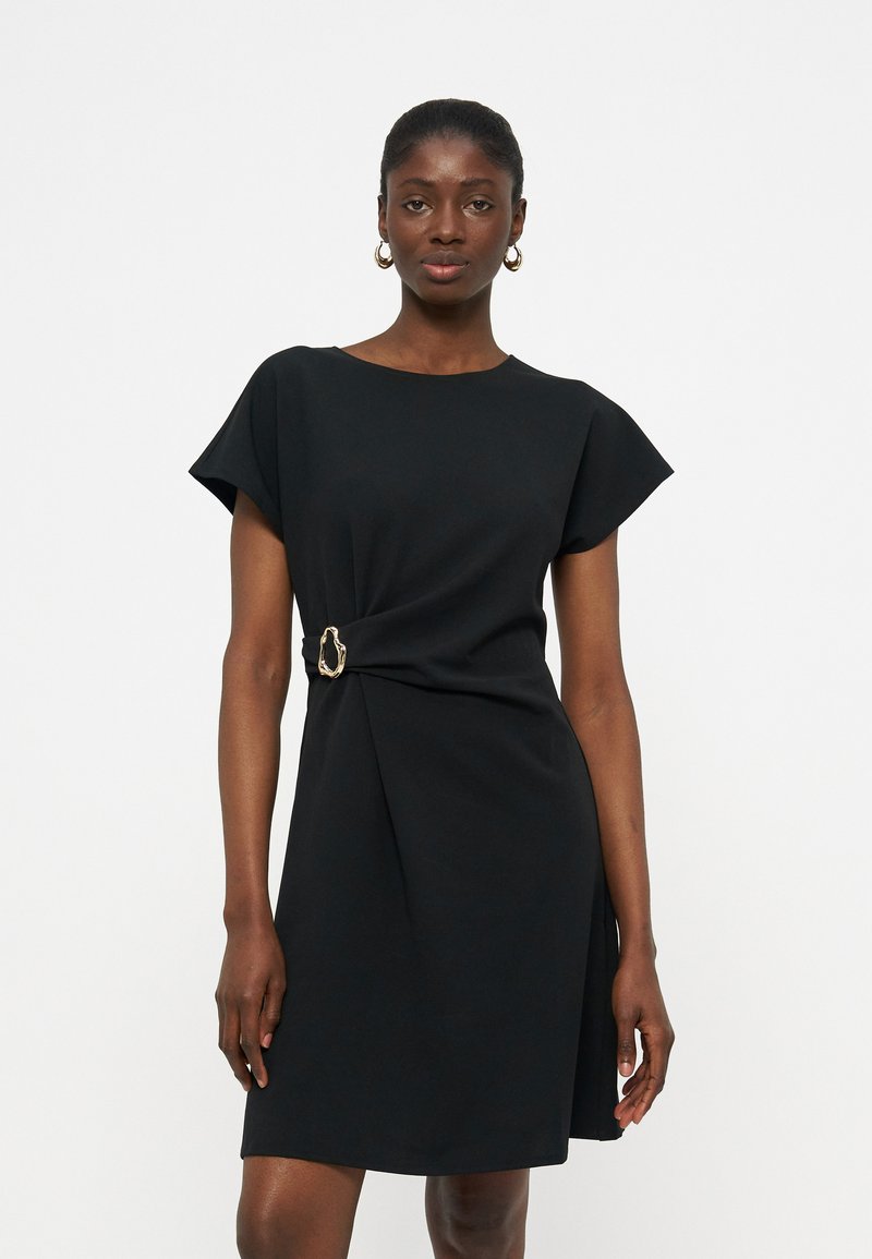 Femme portant une robe noire à manches courtes avec un détail boucle dorée à la taille, debout devant un fond blanc uni.