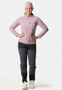 Giacca in pile rosa chiaro con colletto alto e tasca con zip, abbinata a pantaloni cargo grigio scuro con multiple tasche con zip.