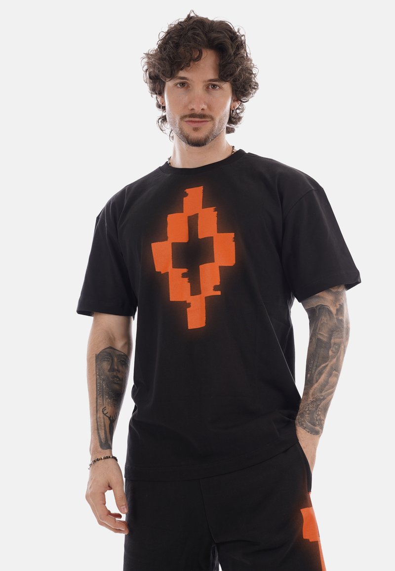 Hombre con cabello rizado y tatuajes que lleva una camiseta negra y pantalones cortos con una gran cruz pixelada naranja en el pecho y la pierna.