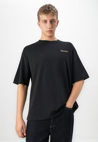 Jeune homme portant un t-shirt noir ample de Timberland avec un logo sur la poitrine et un jean foncé, se tenant devant un fond blanc uni.