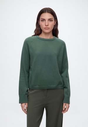 Vrouw met lang bruin haar die gouden oorringen draagt, een groene trui met lange mouwen en donkergroene broek, staand tegen een effen lichte achtergrond.