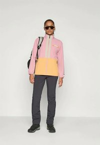 Color-blocked jacket with pink and yellow sections, featuring a high collar, zip pockets, and gray accents. Paired with dark pants and boots.