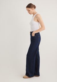 Wide-leg denim jeans in dark blue with subtle stitching, featuring a high waist and two side pockets, paired with a white tank top.