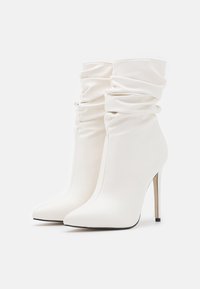 Bottes blanches à talons hauts et cheville avec tissu froncé, bouts pointus et design élégant. Texture brillante notable et talon aigu fin.