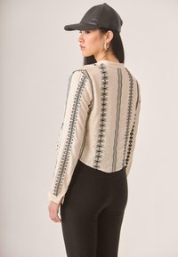 Top corto beige con patrón bordado circular y geométrico en negro, mangas largas y una textura suave. Combinado con pantalones negros.