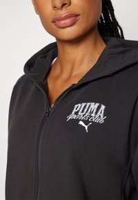Sudadera con cremallera negra de textura suave, con un logo bordado y detalles en blanco. Incluye capucha y cierre frontal de cremallera.