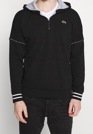 Hombre con sudadera negra con capucha y media cremallera, bolsillo tipo canguro, puños a rayas y pequeño logo verde de cocodrilo en el pecho.