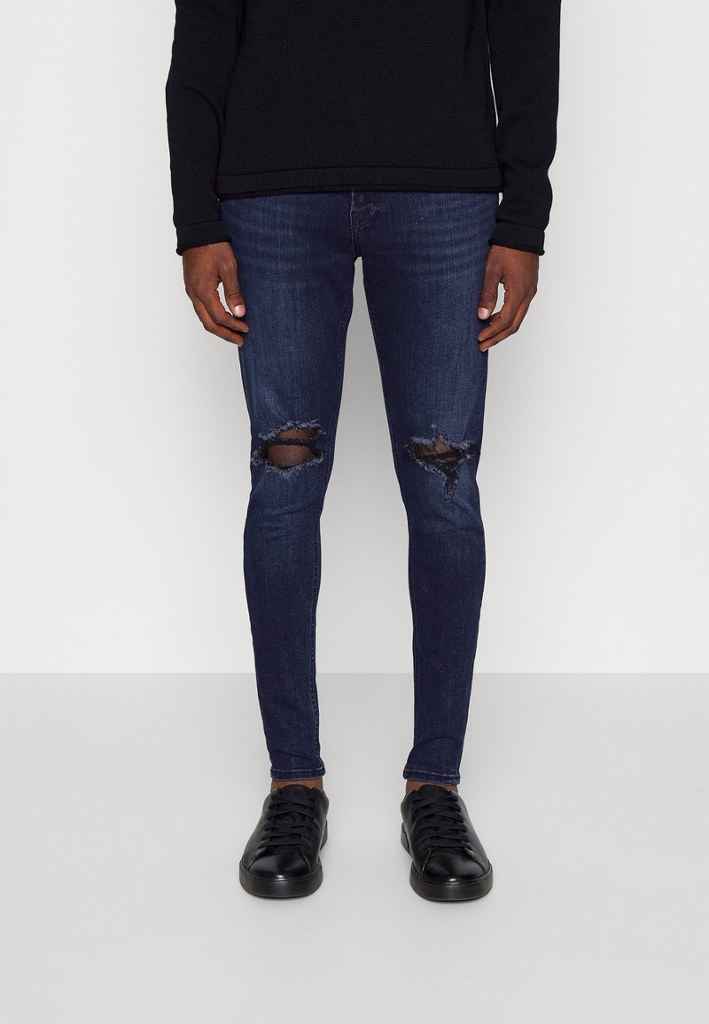 Jean justo escuros de lavagem média com rasgos nos joelhos, combinados com ténis pretos de cano baixo, usados com uma blusa preta de manga longa.