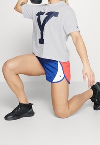Vaaleanharmaa lyhythihainen paita, jossa on suuri navy sininen "Y"-logo, yhdistettynä sinisiin shortsit, joissa on punaisia ja valkoisia yksityiskohtia, sekä mustat urheilukengät.