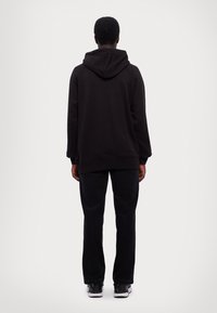 Felpa nera con cappuccio regolabile, vestibilità rilassata, polsini a coste, indossata con jeans scuri e sneakers. Cuciture visibili, tessuto semplice, senza motivi.