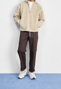 Beige fleece-pullover med kvartdragkedja, med sidofickor och dragsko i nederkant, i kombination med mörkbruna byxor och vita sneakers.