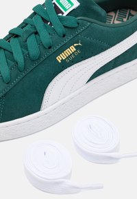 Zelené semišové tenisky s bílým koženým pruhem, s zlatým označením. Obsahuje dvě bílé tkaničky uspořádané do spirály v blízkosti.