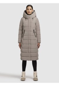 Donna che indossa un lungo cappotto imbottito taupe con un grande cappuccio, leggings neri e stivali beige con lacci, in piedi contro uno sfondo chiaro e semplice.