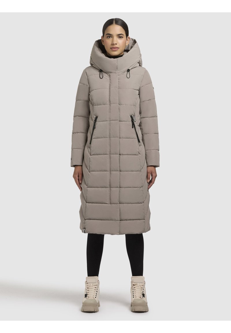Donna che indossa un lungo cappotto imbottito taupe con un grande cappuccio, leggings neri e stivali beige con lacci, in piedi contro uno sfondo chiaro e semplice.