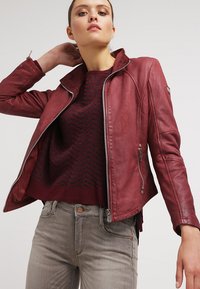 Giubbotto da motociclista in pelle rossa con chiusura a zip, dotato di colletto rialzato, cerniere laterali e cuciture dettagliate. Indossato sopra un maglione bordeaux.