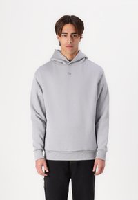 Felpa grigia chiaro realizzata in tessuto morbido, con cappuccio regolabile, polsini a coste e un piccolo logo sul petto. Indossata con pantaloni scuri.