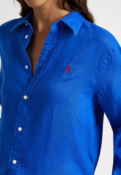 Μπλε λινό πουκάμισο με κουμπιά, γιακά, λευκά κουμπιά και κόκκινο κεντημένο λογότυπο στο αριστερό στήθος. Λεία υφή, άνετη εφαρμογή.