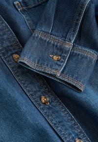 Denimová košile v tmavě modré barvě, s béžovým prošíváním a zapínáním na knoflíky. Obsahuje ohnutý manžetový detail s knoflíkem.