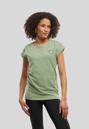 Femme aux cheveux bouclés portant un t-shirt vert clair avec le contour noir d'un petit visage de chien sur la poitrine, associé à un pantalon noir, debout devant un fond gris.