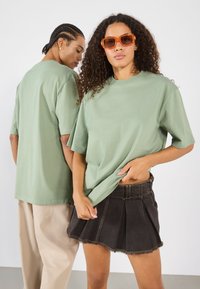 Grønn oversized t-skjorte i myk stoff, kombinert med et svart denimskjørt med slitt kant og folder. Begge modellene står med ryggen og ansiktet vendt fremover.