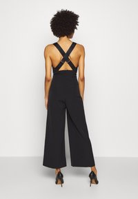 Combinaison noire à jambes larges, dotée de bretelles croisées dans le dos et d'une fermeture éclair. Tissu lisse avec des accents de logo sur les bretelles.