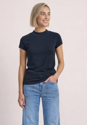 Frau mit kurzen blonden Haaren, die ein dunkelblaues kurzärmliges Hemd und hellblaue Jeans trägt, lächelnd und zur Seite schauend.