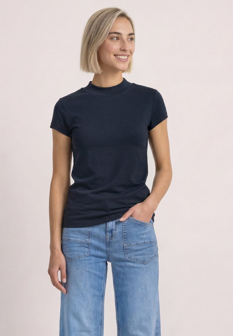 Femme aux cheveux blonds courts portant un t-shirt bleu marine à manches courtes et un jean bleu clair, souriante et regardant sur le côté.