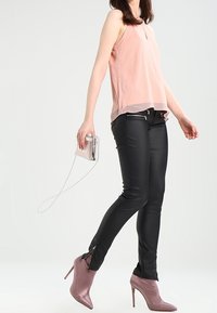 Light pink sleeveless top with pleated texture, paired with black slim-fit pants featuring zipper accents and side slits, worn with pink heeled shoes.
