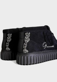 Schwarze Wildleder-Stiefeletten mit strukturiertem Finish. Sie verfügen über eine klobige schwarze Sohle, gestickte Verzierungen und ein seitliches Logo.
