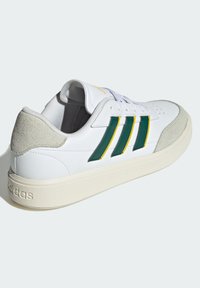 Bijela tenisica s zelenim i žutim prugama, teksturiranim nubukakima na peti i glatkom gumiranom potplatu s Adidas logotipom.