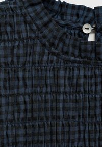 Temno modra karirana tkanina z nabranim vzorcem. Ima nabran ovratnik in gumb na hrbtni strani.