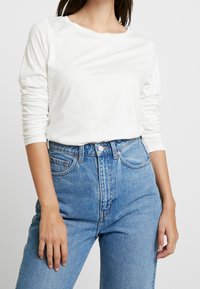 Langärmliges weißes Baumwollhemd mit Rundhalsausschnitt, kombiniert mit hochgeschnittenen blauen Jeans aus Denim, die klassische Taschen und einen verwaschenen Look aufweisen.